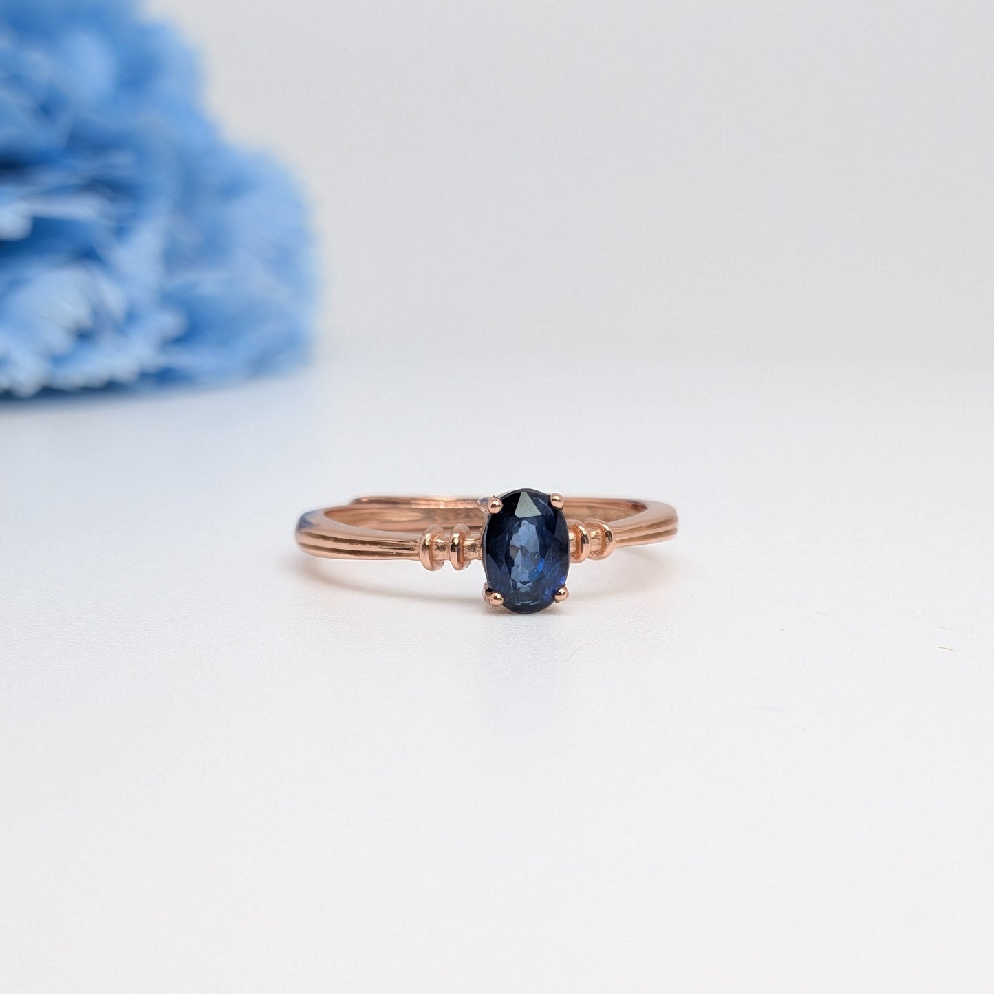 A delicate rose gold ring with a small oval dark blue gemstone, displayed on a clean white surface with a soft-focus blue flower in the background.