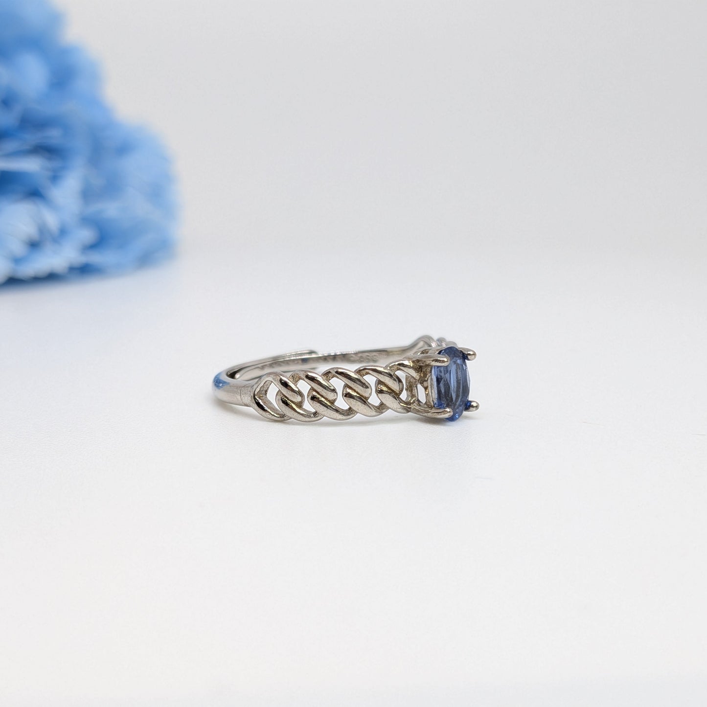 A silver ring with a braided band and a rectangular blue gemstone, photographed on a white background with a soft-focus blue flower in the upper left corner.