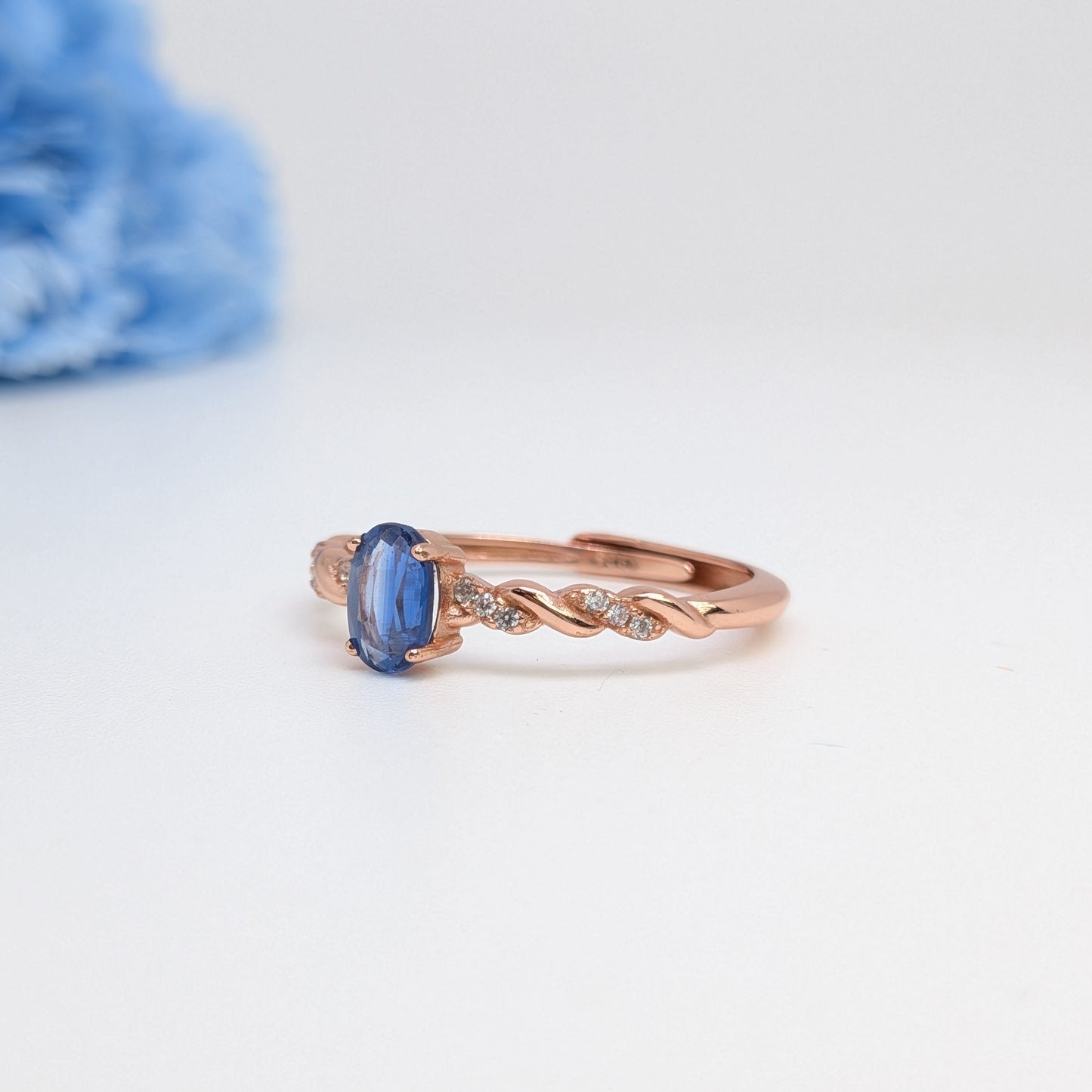 A rose gold ring with a blue oval gemstone and small clear accent stones, displayed on a clean white surface with a soft-focus blue flower in the background.