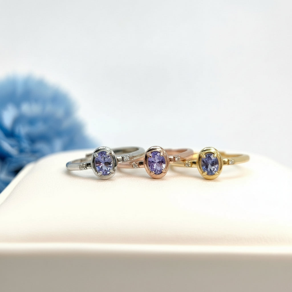 Three gold rings with purple gemstones on a light surface with a blurred blue flower in the background.