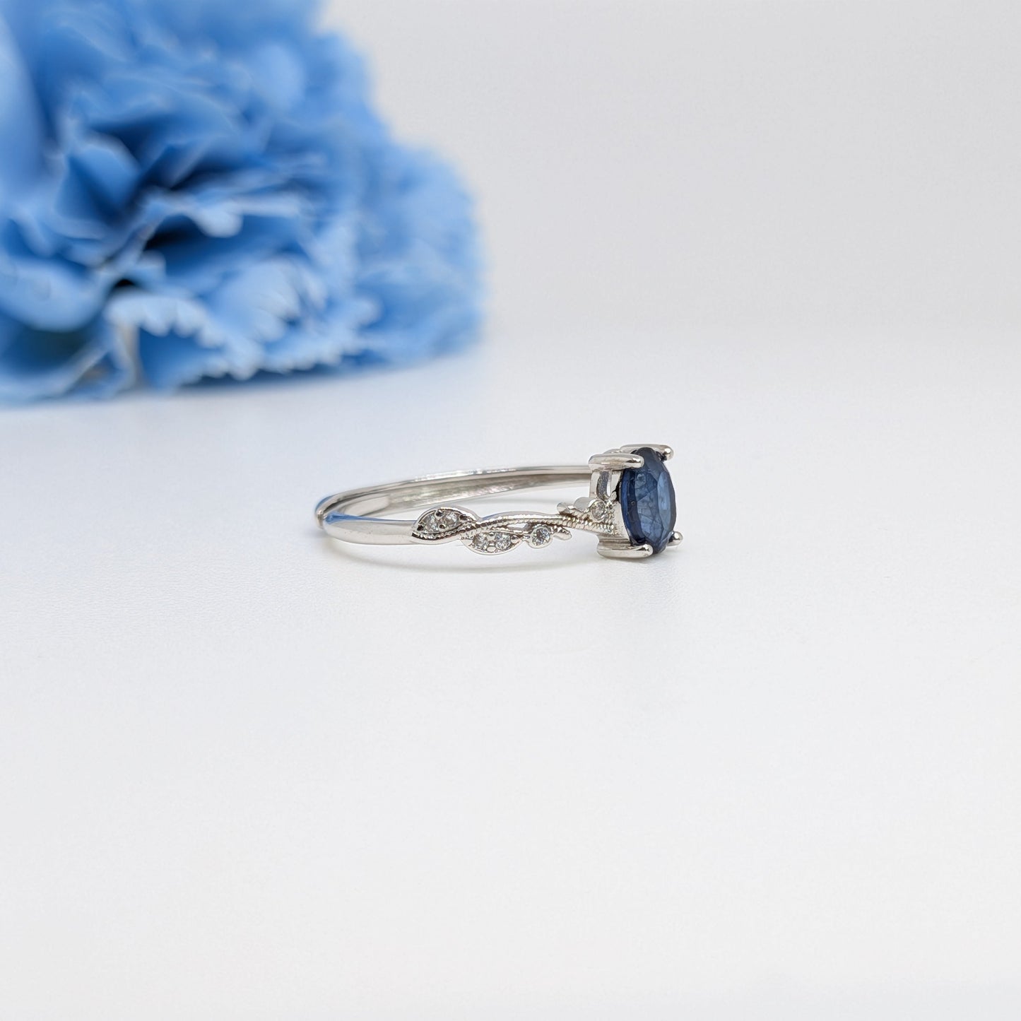 A silver ring with a marquise-cut dark blue sapphire and small pavé diamonds on the band, displayed on a clean white surface with a blurred blue flower in the background.