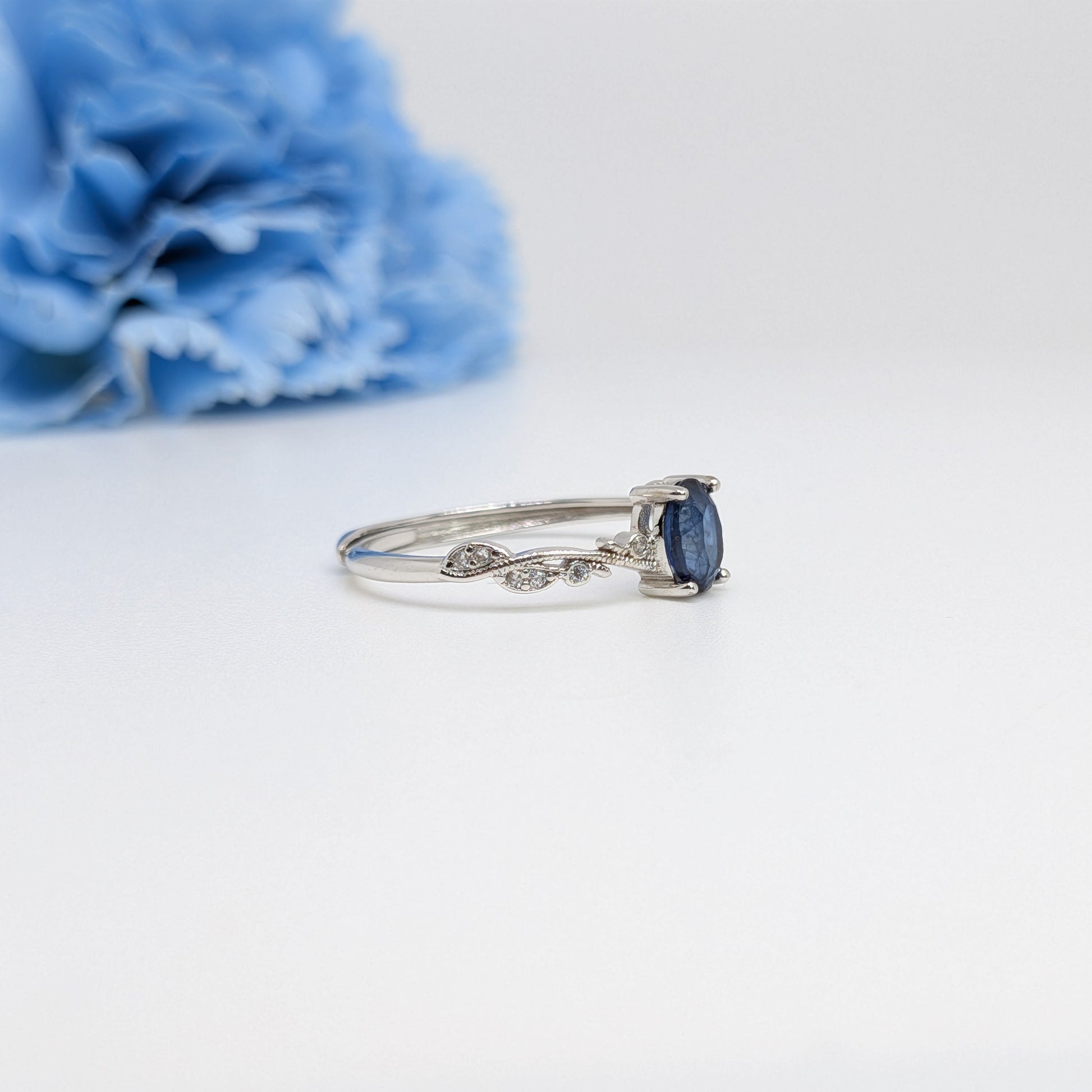 A silver ring with a marquise-cut dark blue sapphire and small pavé diamonds on the band, displayed on a clean white surface with a blurred blue flower in the background.