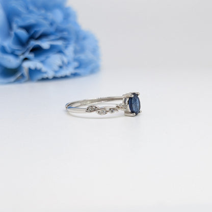 A silver ring with a marquise-cut dark blue sapphire and small pavé diamonds on the band, displayed on a clean white surface with a blurred blue flower in the background.