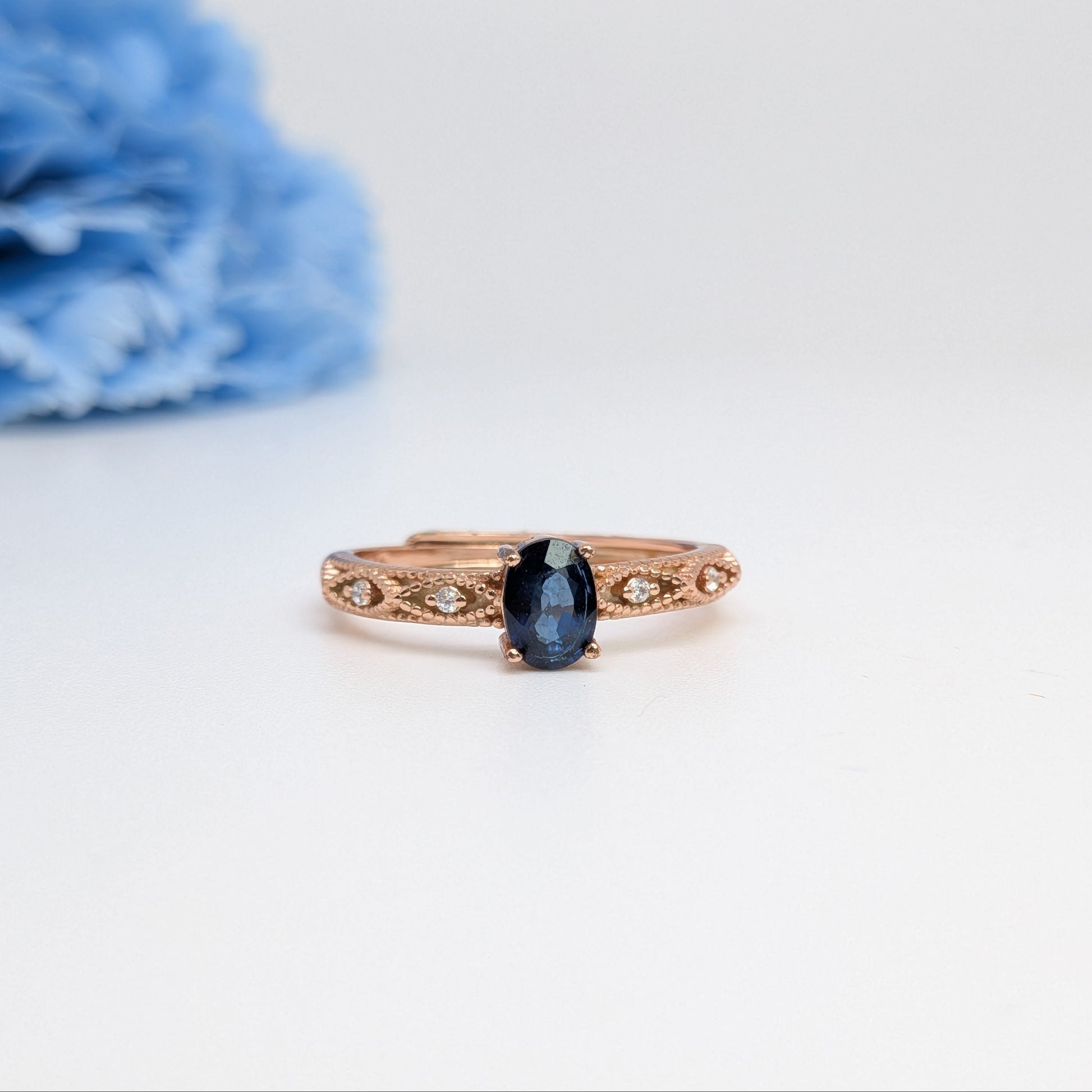 A rose gold ring with an oval-cut dark blue sapphire center stone and small pavé-set accent stones along the band, displayed on a clean white surface with a soft-focus blue flower in the background.