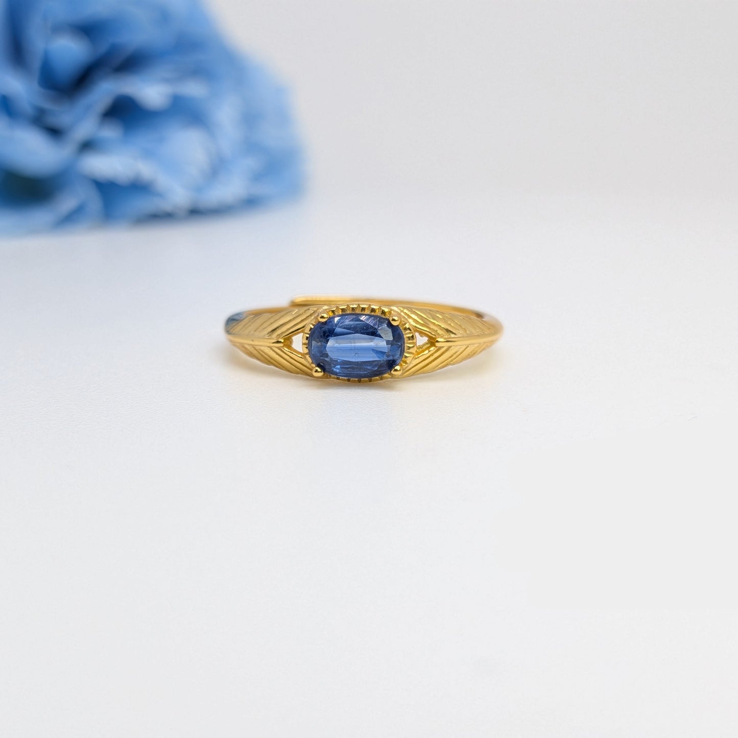A yellow gold ring with an oval-cut blue gemstone in the center, flanked by small clear stones and decorative gold detailing, set against a white background with a blurred blue flower in the upper left.