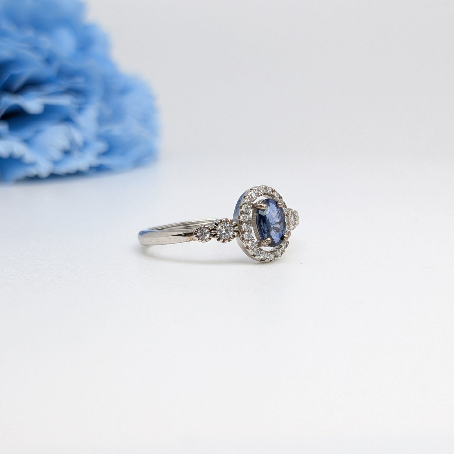 A silver-toned ring featuring an oval blue sapphire center stone surrounded by a halo of small clear stones, with a slim matching band, displayed on a white surface with a soft-focus blue flower in the background.