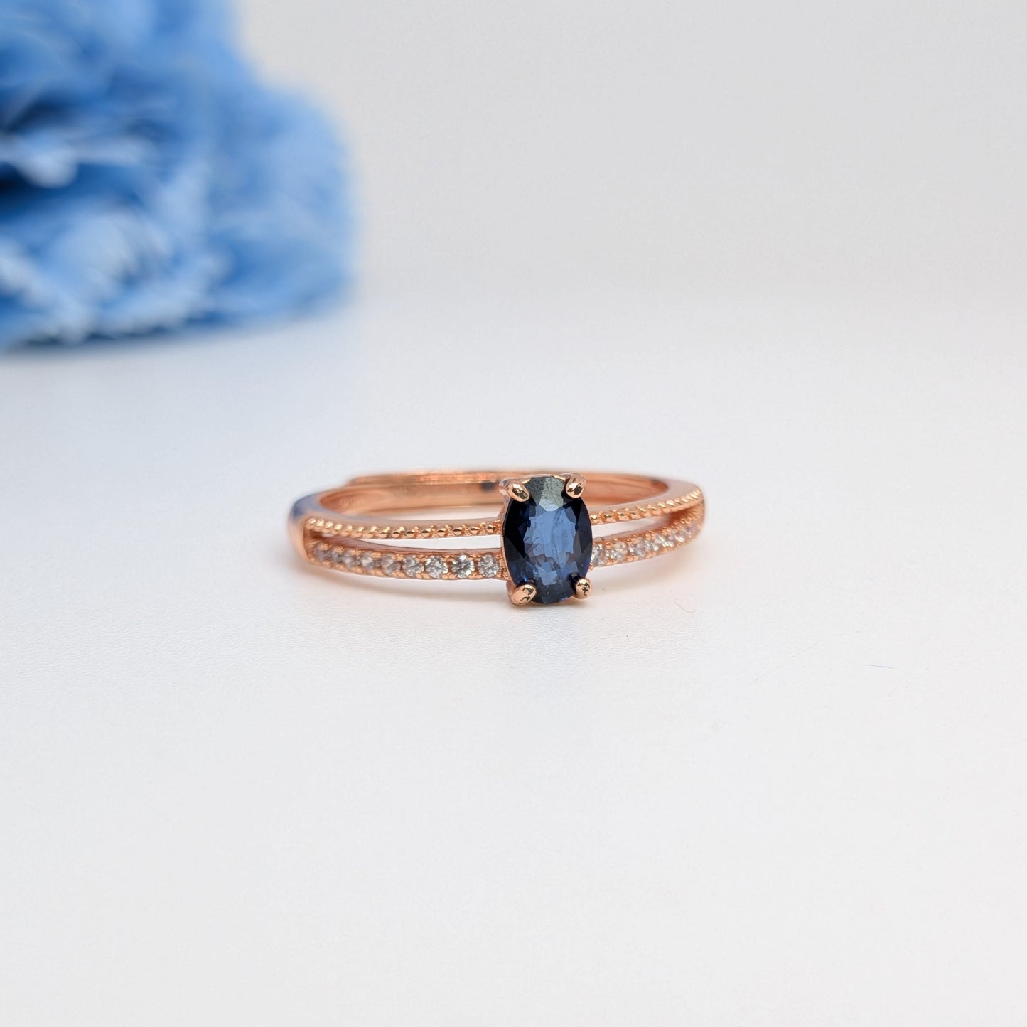 A rose gold ring featuring an oval-cut dark blue sapphire center stone and a pavé band with small clear stones, set against a white background with a soft-focus blue flower in the upper left.