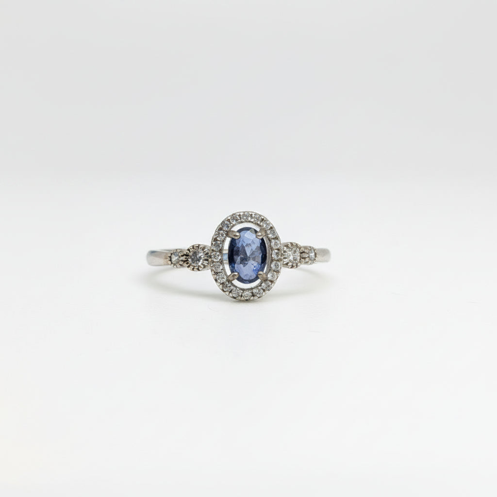 An oval-cut blue sapphire ring set in a silver-colored halo with small accent stones, displayed on a clean white surface with a soft-focus blue flower in the background.