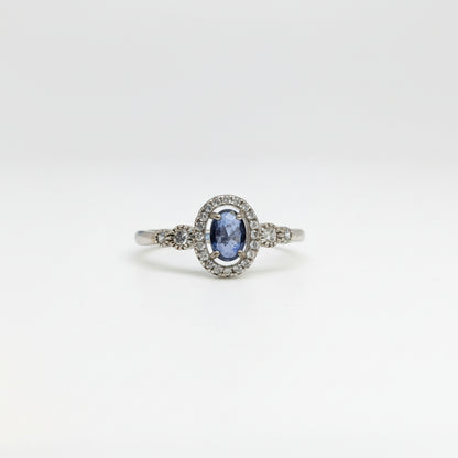 An oval-cut blue sapphire ring set in a silver-colored halo with small accent stones, displayed on a clean white surface with a soft-focus blue flower in the background.