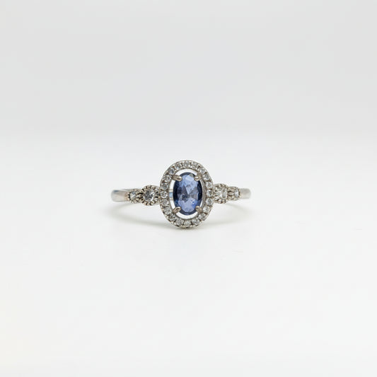 An oval-cut blue sapphire ring set in a silver-colored halo with small accent stones, displayed on a clean white surface with a soft-focus blue flower in the background.