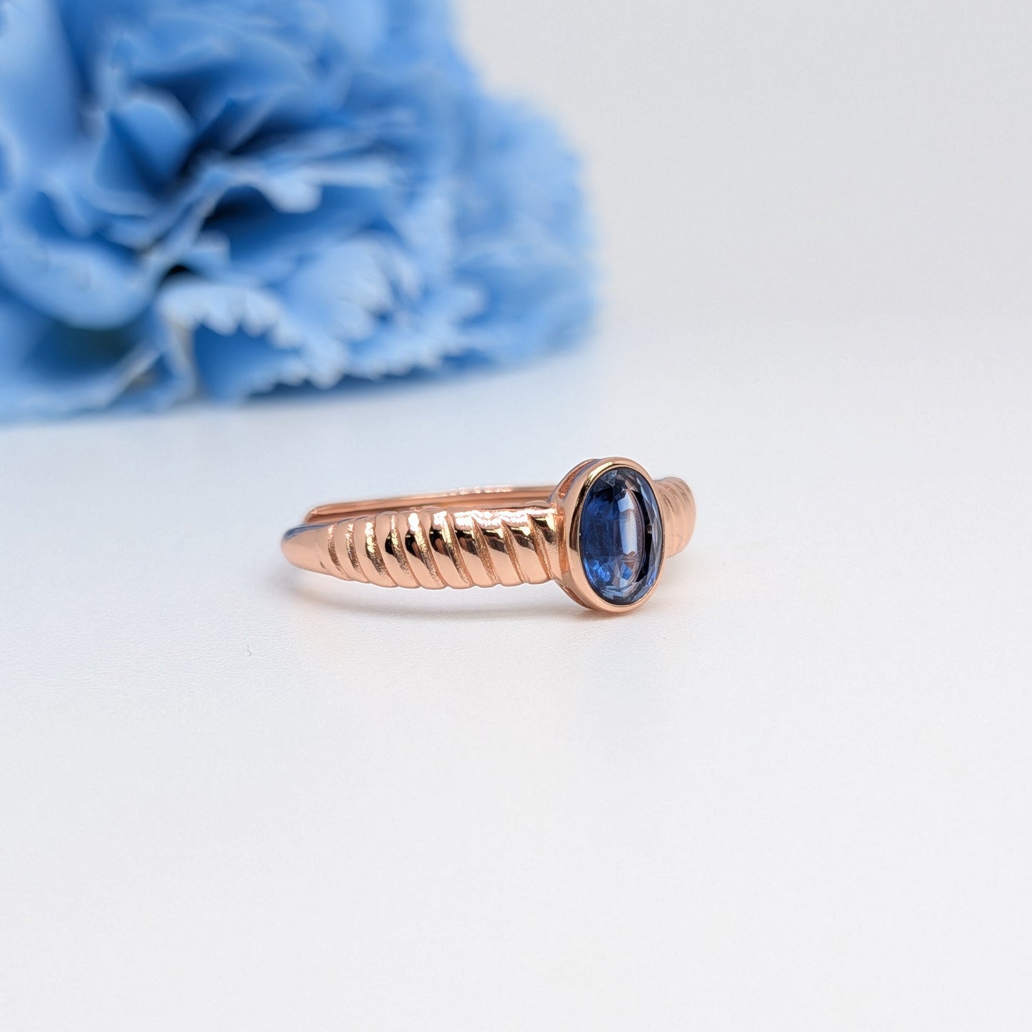 A rose gold ring with a textured band and an oval blue gemstone, photographed on a clean white surface with a soft-focus blue flower in the background.