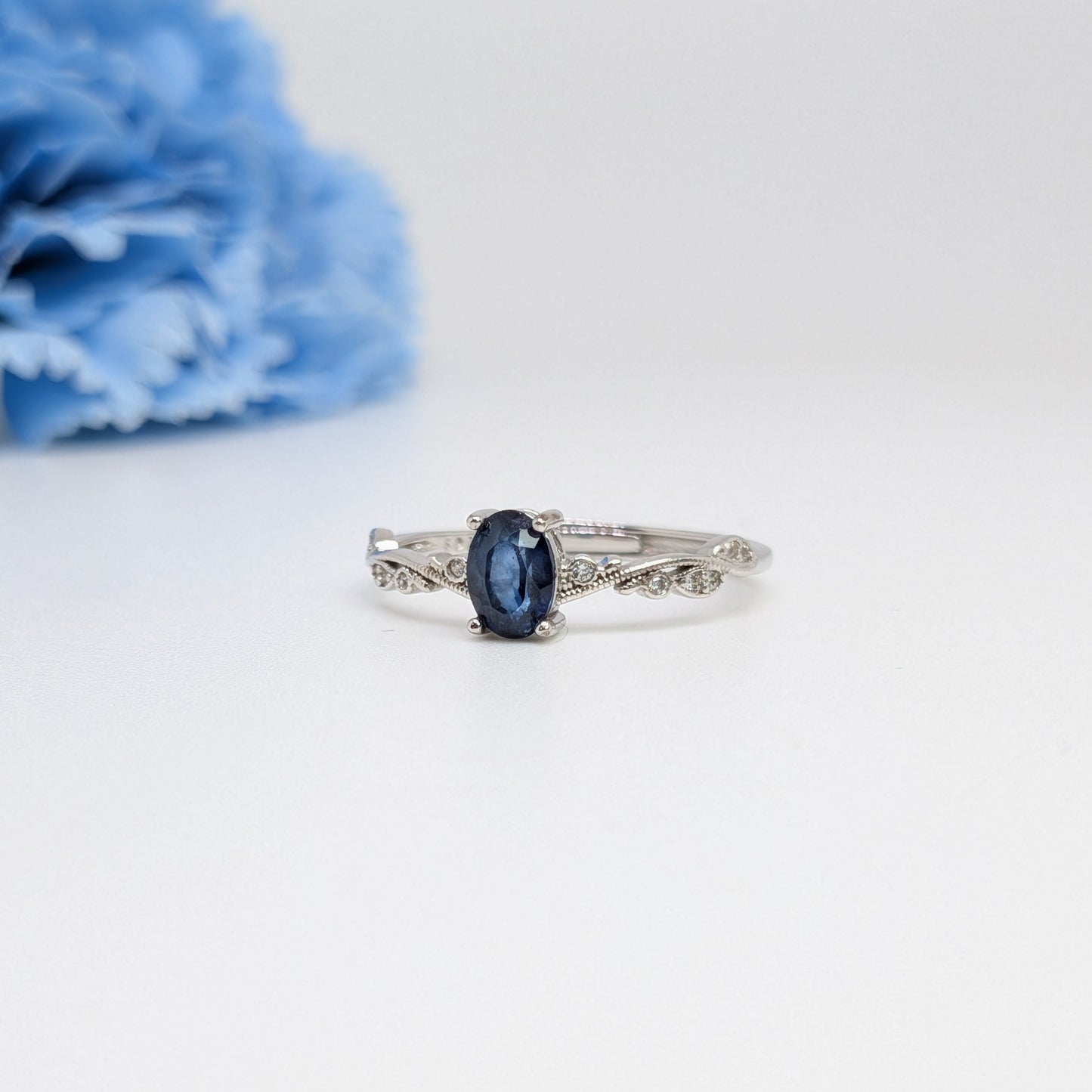 A silver ring with an oval-cut dark blue gemstone, displayed on a clean white surface with a blurred light blue flower in the background.