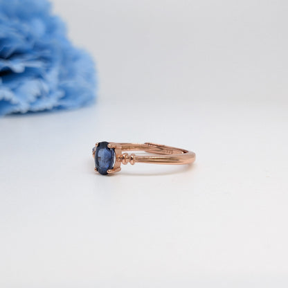 A rose gold ring with a small oval blue gemstone, photographed on a clean white surface with a soft-focus blue flower in the background.