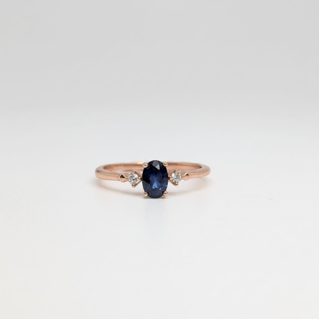 A rose gold ring featuring a central oval-cut dark blue sapphire gemstone, accented by two small round-cut clear stones on either side of the band, set against a white background with a soft-focus blue flower in the upper left corner.