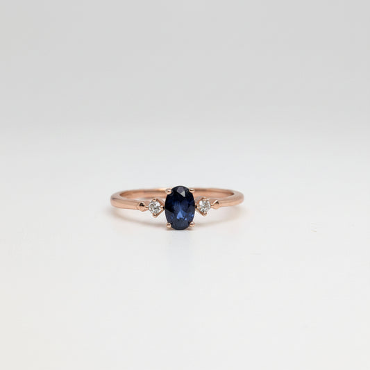 A rose gold ring featuring a central oval-cut dark blue sapphire gemstone, accented by two small round-cut clear stones on either side of the band, set against a white background with a soft-focus blue flower in the upper left corner.