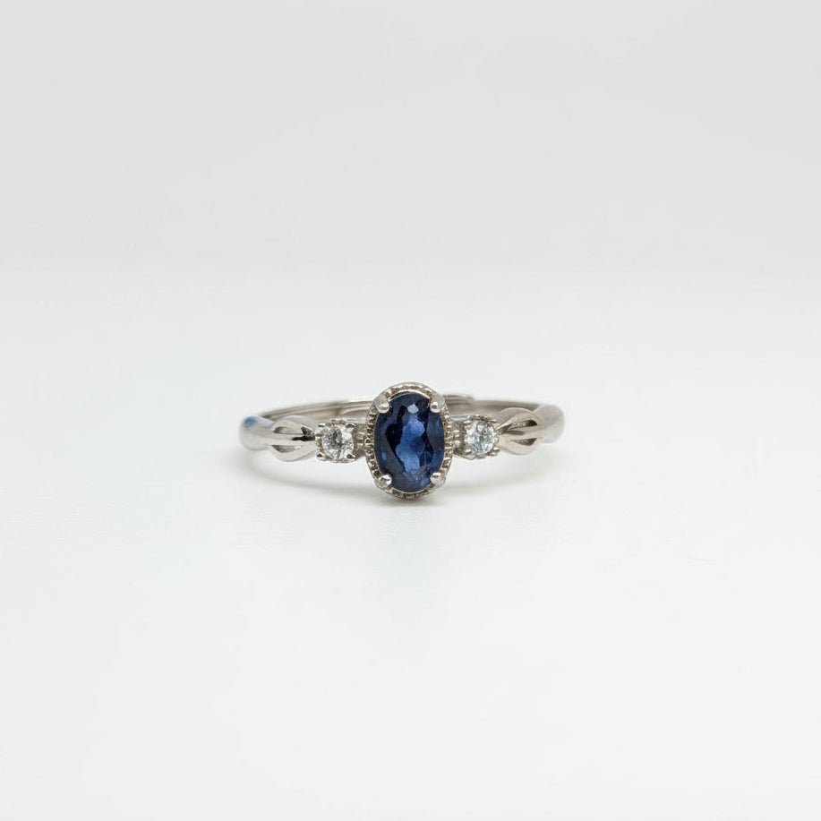 A silver ring featuring an oval-cut dark blue sapphire center stone and small clear side stones, displayed on a clean white surface with a soft-focus blue flower in the background.