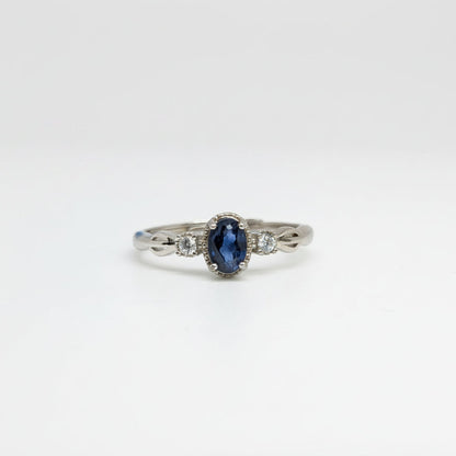 A silver ring featuring an oval-cut dark blue sapphire center stone and small clear side stones, displayed on a clean white surface with a soft-focus blue flower in the background.
