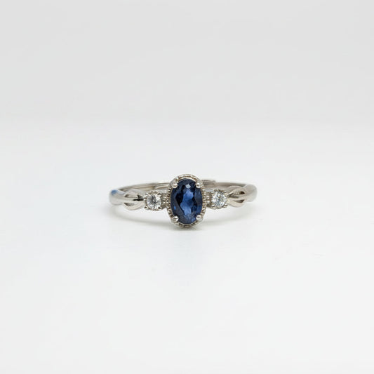 A silver ring featuring an oval-cut dark blue sapphire center stone and small clear side stones, displayed on a clean white surface with a soft-focus blue flower in the background.