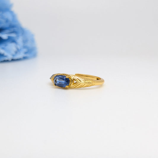 A gold ring with a marquise-cut blue gemstone and a textured leaf-like setting, displayed on a clean white surface with a soft-focus blue flower in the background.
