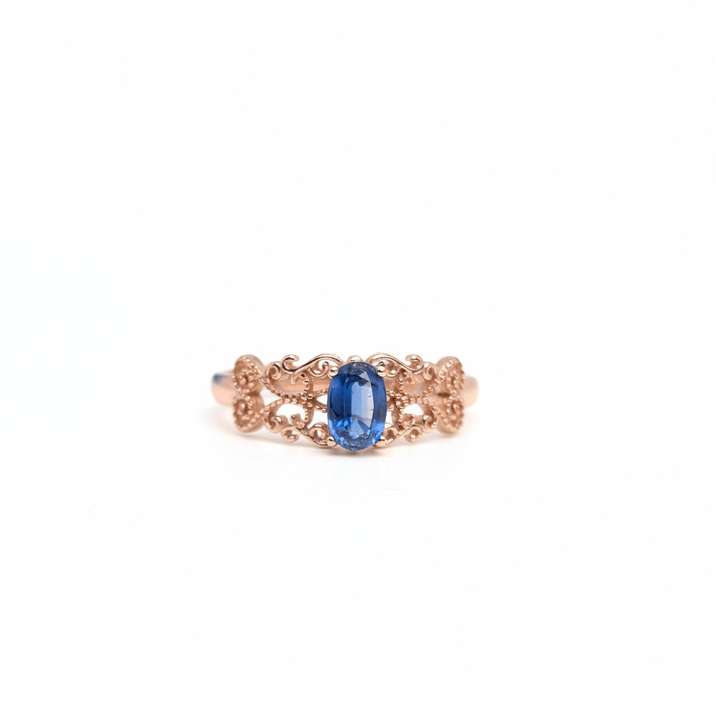 A delicate rose gold ring with an oval-cut blue sapphire center stone, set against a clean white background with a soft-focus blue flower in the upper left corner.