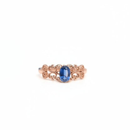 A delicate rose gold ring with an oval-cut blue sapphire center stone, set against a clean white background with a soft-focus blue flower in the upper left corner.