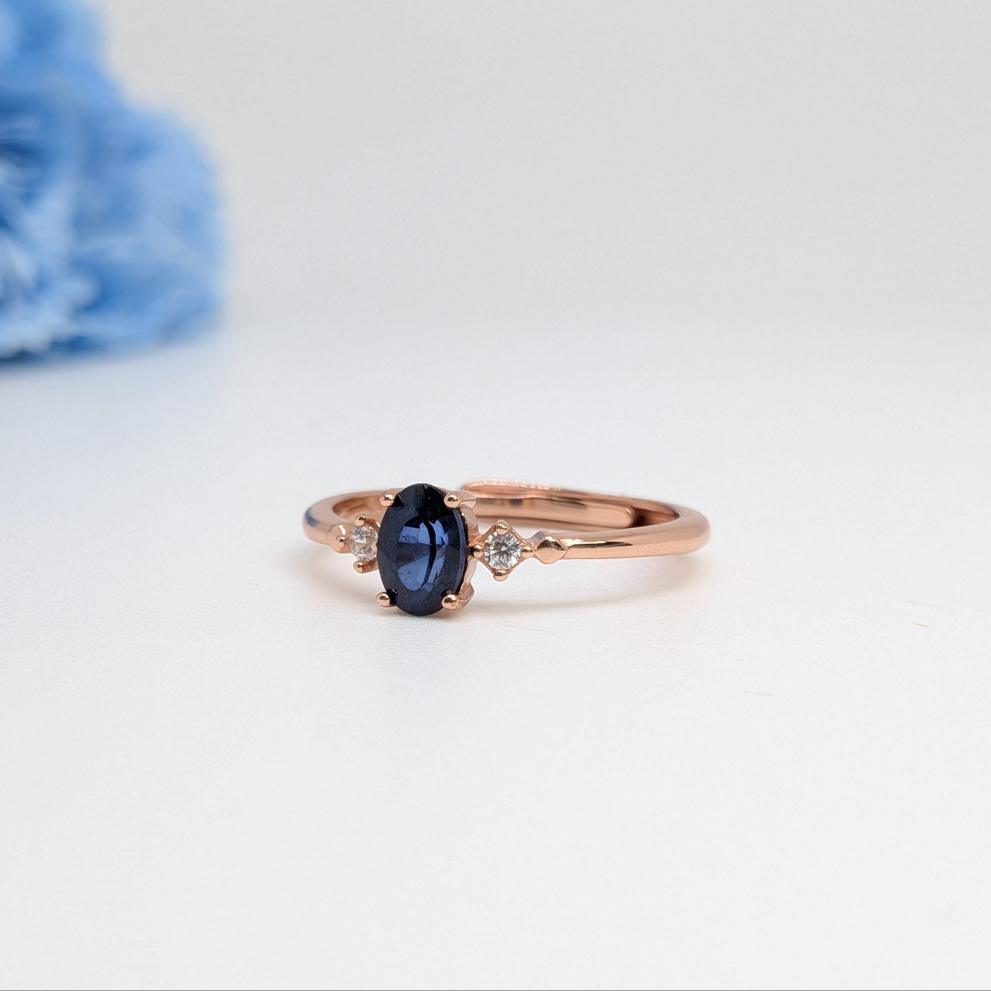A rose gold ring with an oval dark blue sapphire center stone and a small clear diamond accent on each side, displayed on a clean white surface with a soft-focus blue flower in the background.