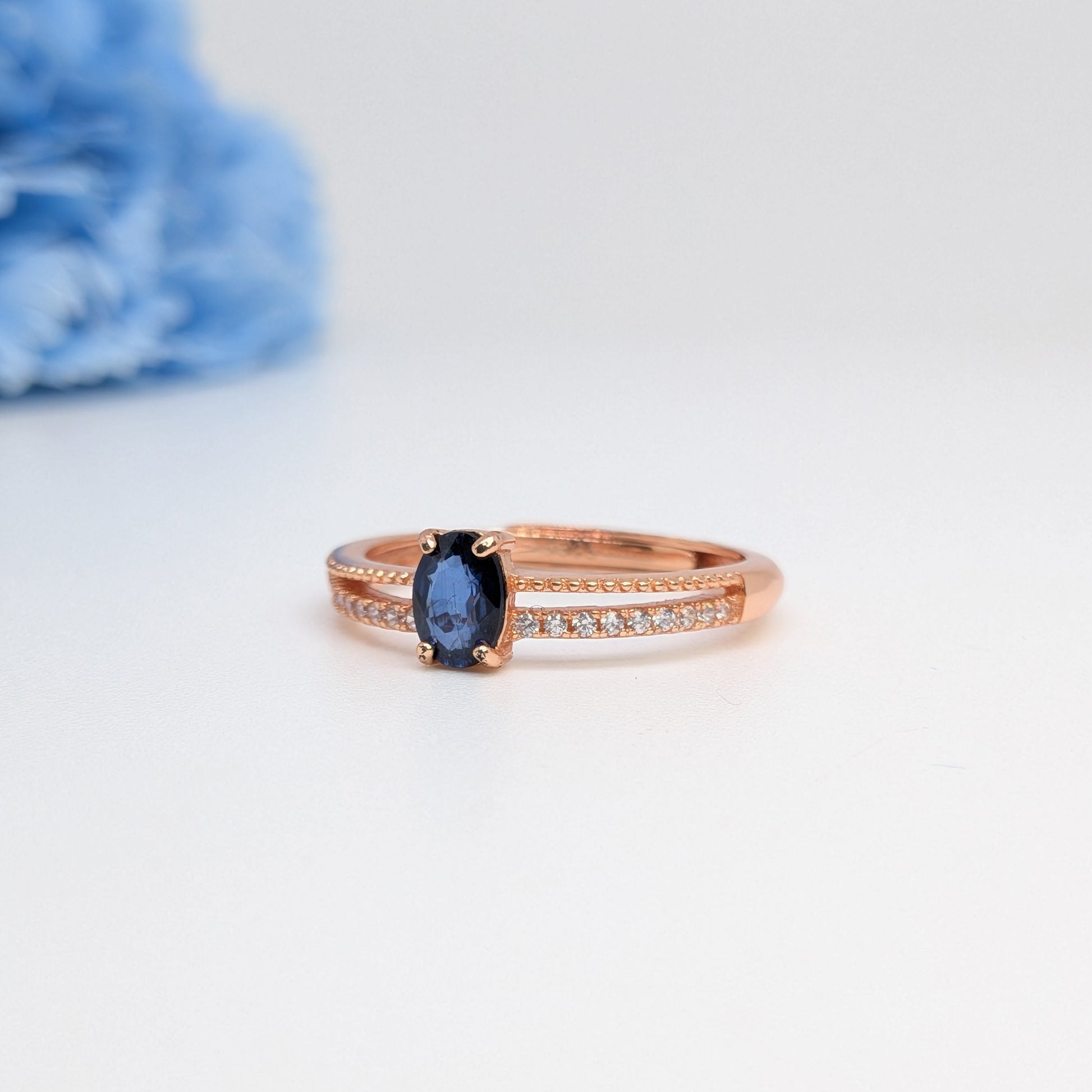 A rose gold ring with a central oval-cut dark blue gemstone and a pavé band of small clear stones, photographed on a white background with a soft-focus blue flower in the upper left corner.