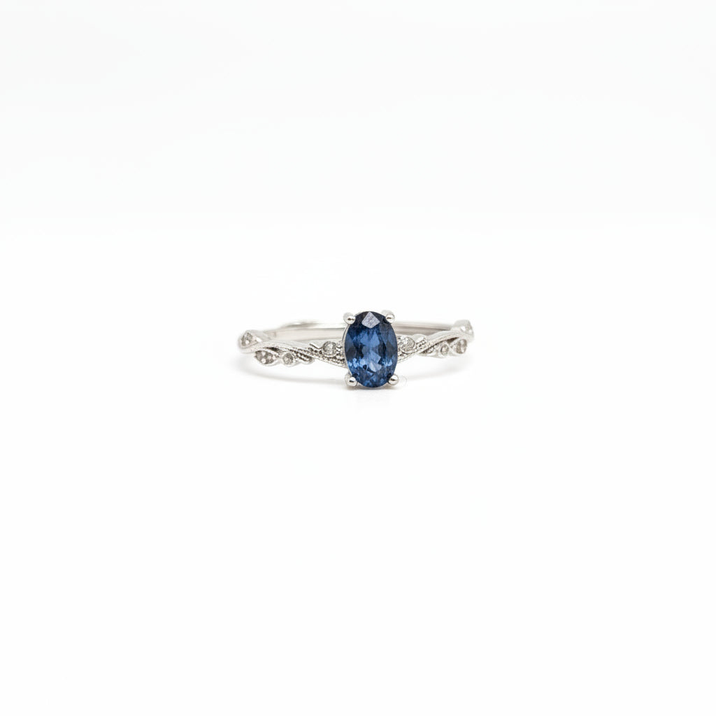 A silver ring with an oval-cut dark blue gemstone, displayed on a clean white surface with a soft-focus light blue flower in the background.