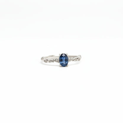 A silver ring with an oval-cut dark blue gemstone, displayed on a clean white surface with a soft-focus light blue flower in the background.