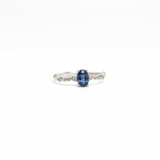 A silver ring with an oval-cut dark blue gemstone, displayed on a clean white surface with a soft-focus light blue flower in the background.