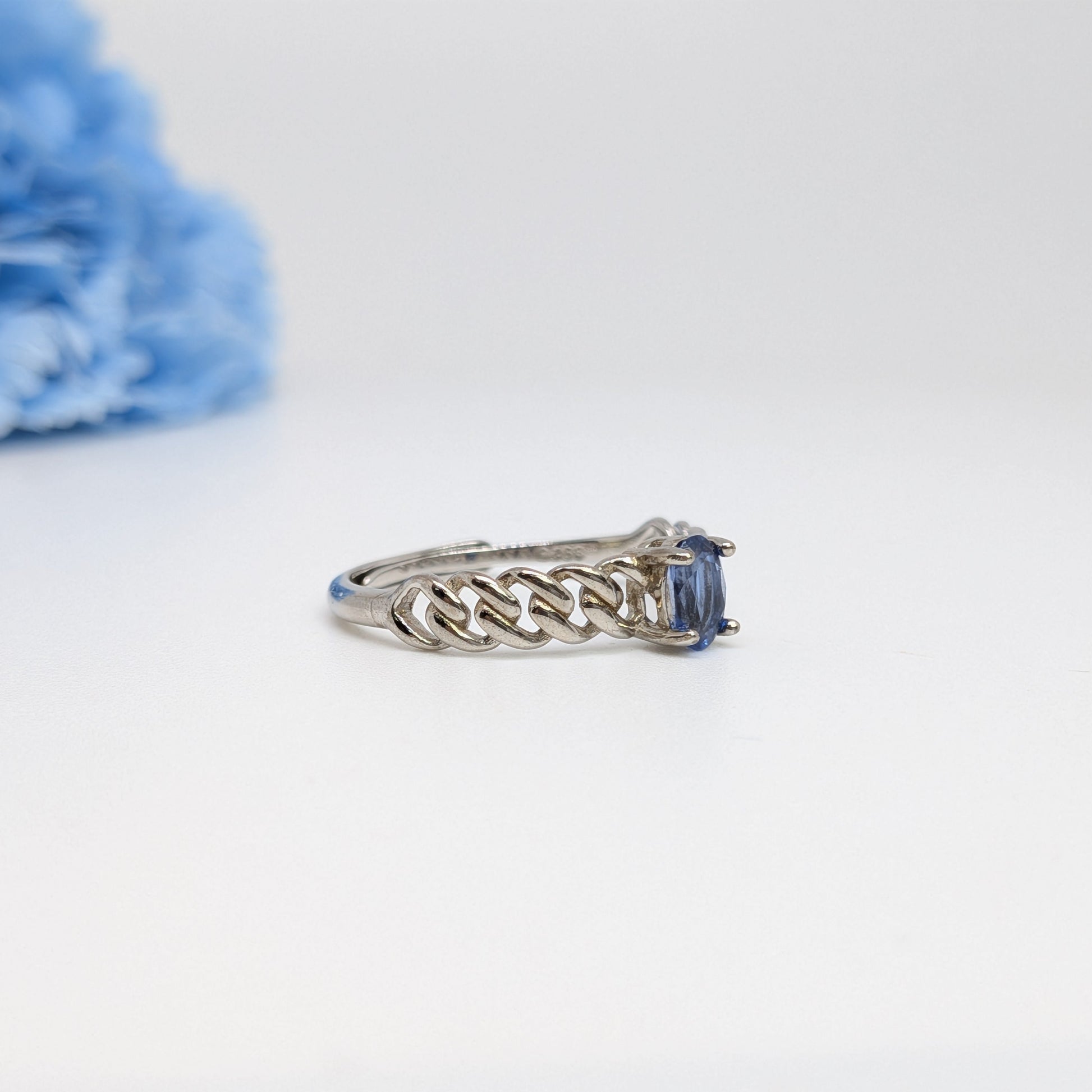 A silver ring with a braided band and a rectangular blue gemstone, photographed on a white background with a soft-focus blue flower in the upper left corner.