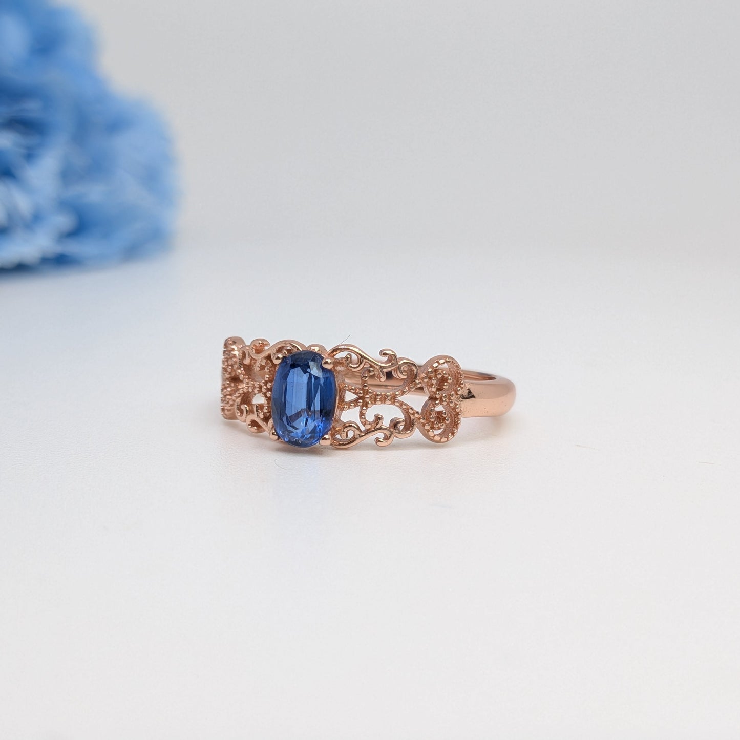 A rose gold ring with an ornate filigree band and a central rectangular blue gemstone, photographed on a white surface with a blurred blue flower in the background.