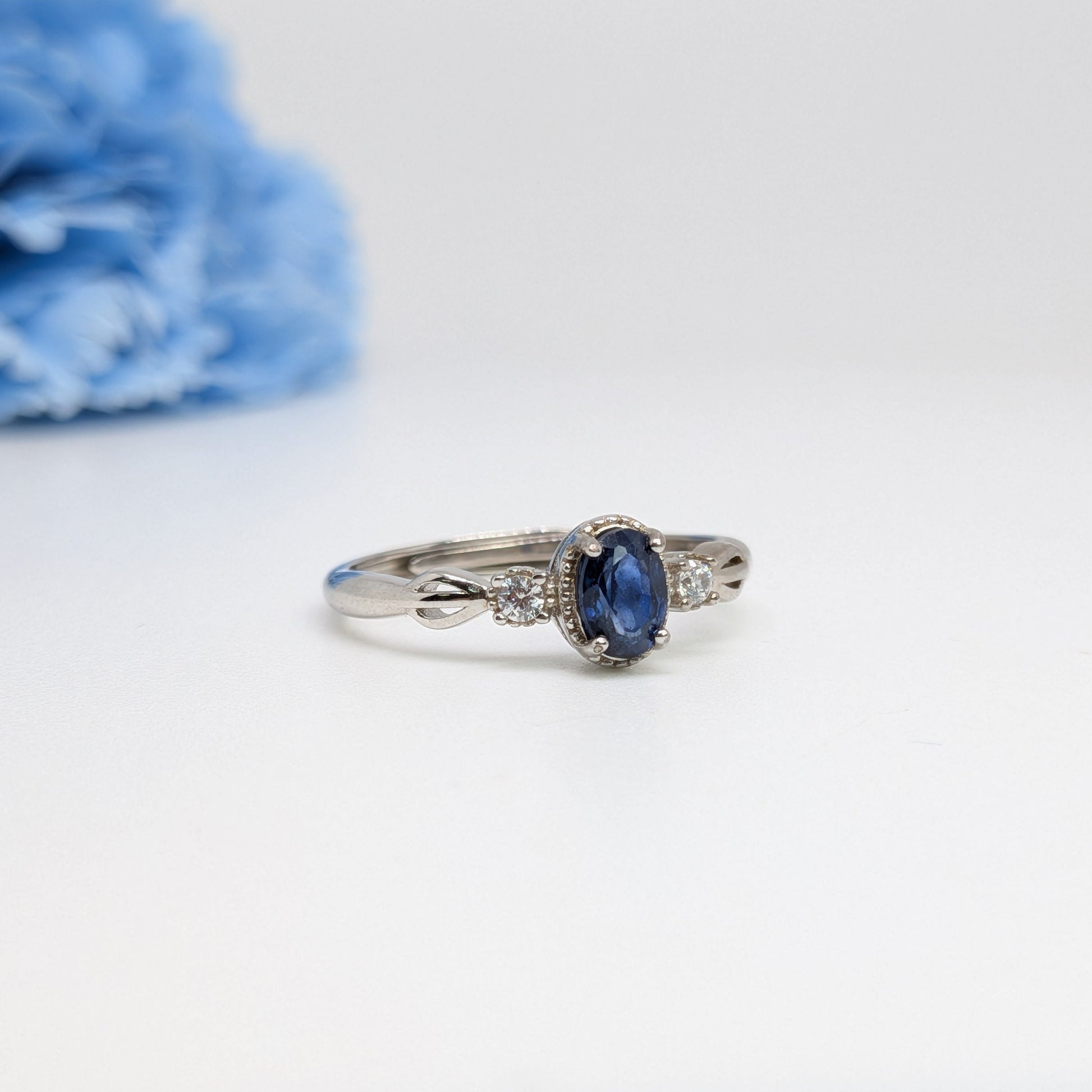 A silver ring featuring an oval-cut dark blue sapphire center stone and small clear accent stones on the band, displayed on a white surface with a soft-focus blue flower in the background.