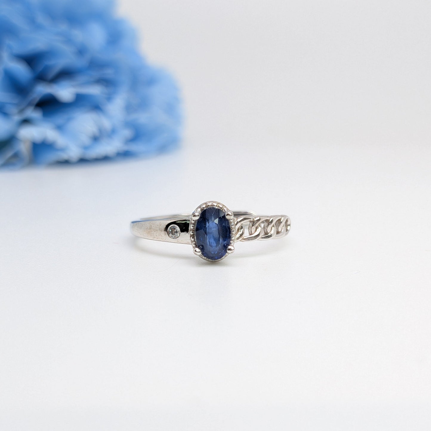 A silver ring with an oval blue gemstone and a decorative chain-link design on the band, displayed on a white surface with a blurred blue flower in the background.