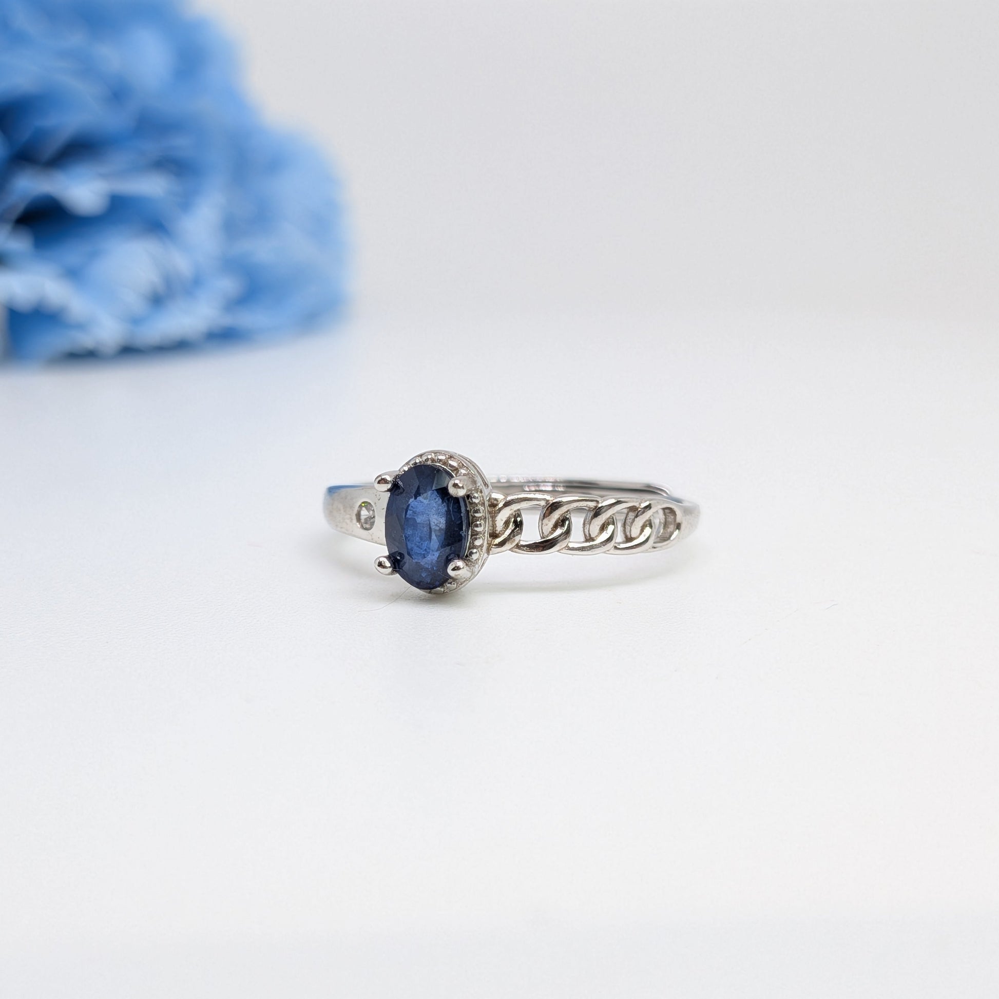 A silver ring with an oval blue gemstone in a decorative setting, displayed on a clean white surface with a soft-focus blue flower in the background.