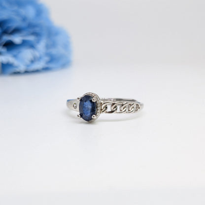 A silver ring with an oval blue gemstone in a decorative setting, displayed on a clean white surface with a soft-focus blue flower in the background.