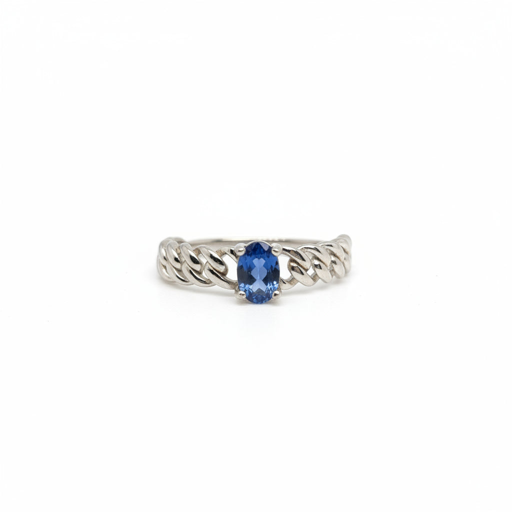 A silver-toned ring with a textured, braided band and a central oval-cut blue gemstone, displayed on a clean white surface with a soft-focus blue flower in the background.