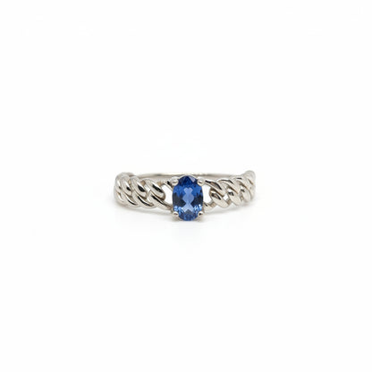 A silver-toned ring with a textured, braided band and a central oval-cut blue gemstone, displayed on a clean white surface with a soft-focus blue flower in the background.