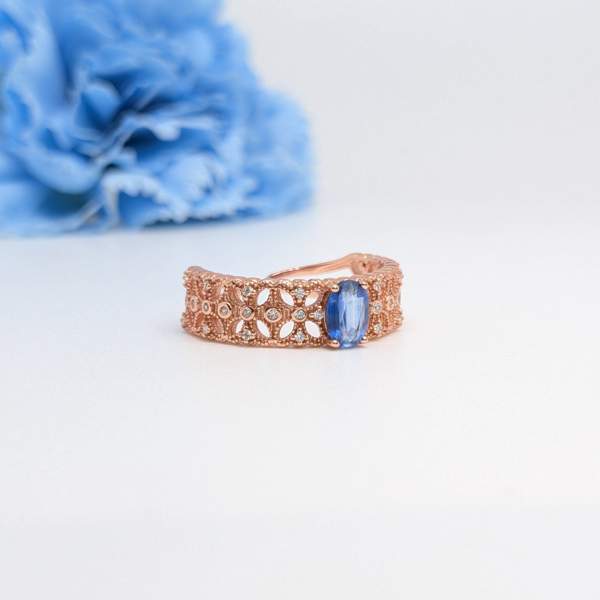 A rose gold ring with an intricate filigree pattern and a rectangular blue gemstone, displayed on a clean white surface with a blurred blue flower in the background.