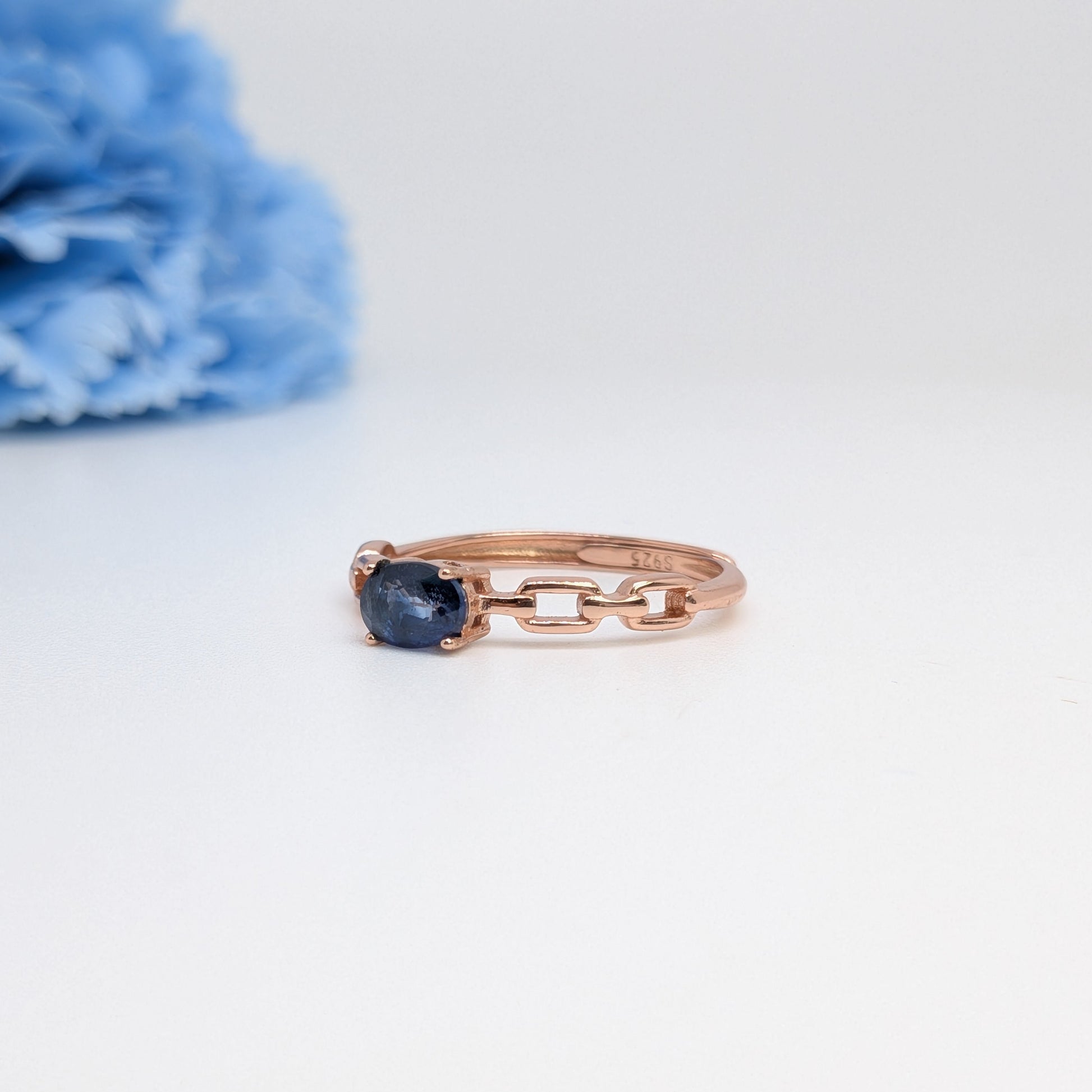 A rose gold ring with a small oval dark blue gemstone and a chain-link design on the band, displayed on a white surface with a blurred blue flower in the background.