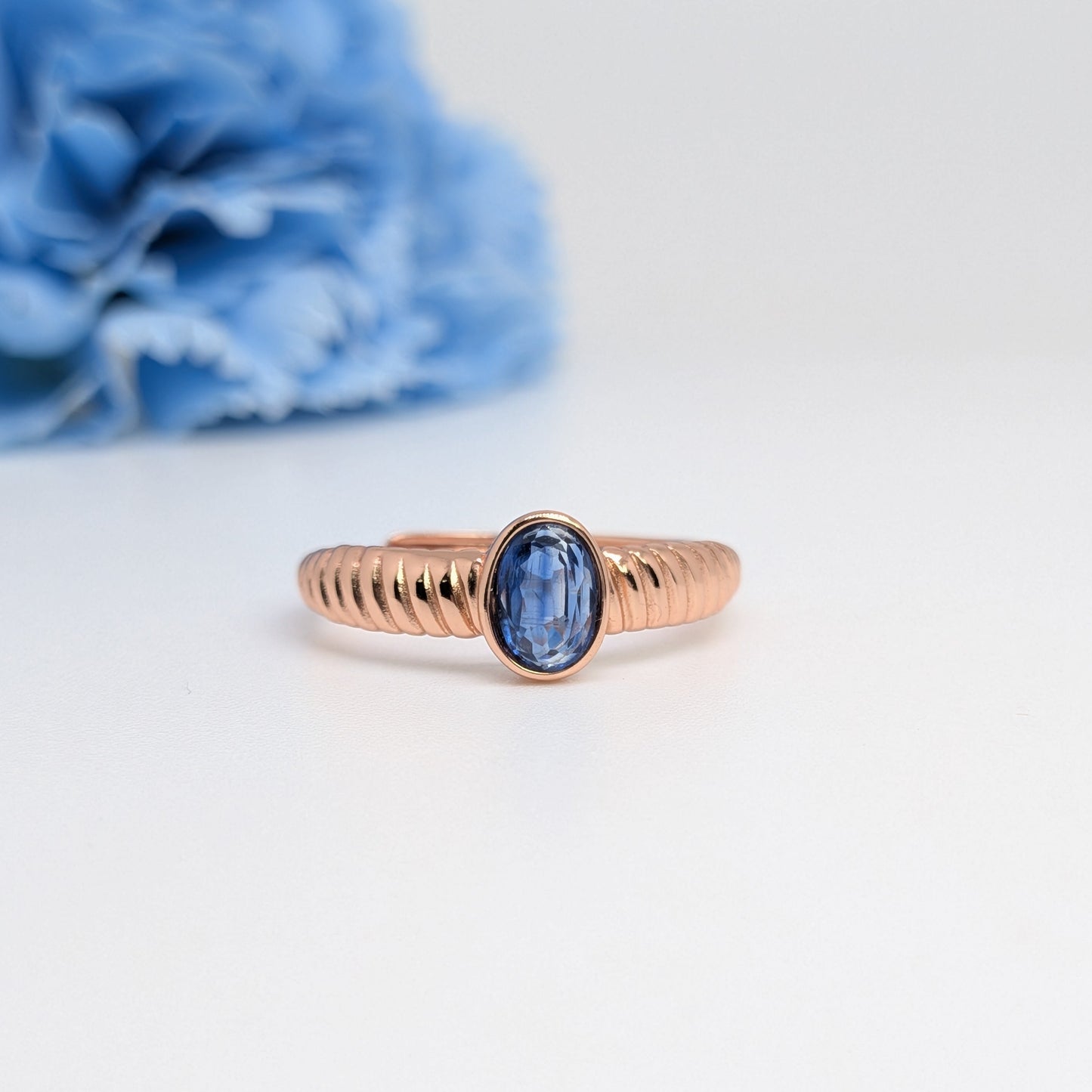 A rose gold ring with a textured band and an oval blue gemstone, displayed on a clean white surface with a blurred blue flower in the background.