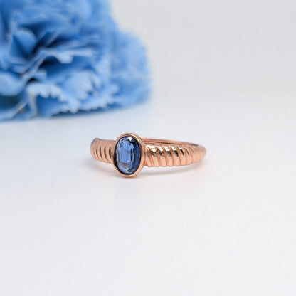 A rose gold ring with a textured band and an oval-cut blue gemstone, displayed on a clean white surface with a blurred blue flower in the background.