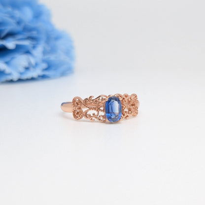 A rose gold ring with an oval-cut blue sapphire center stone, displayed on a clean white surface with a soft-focus blue flower in the background.