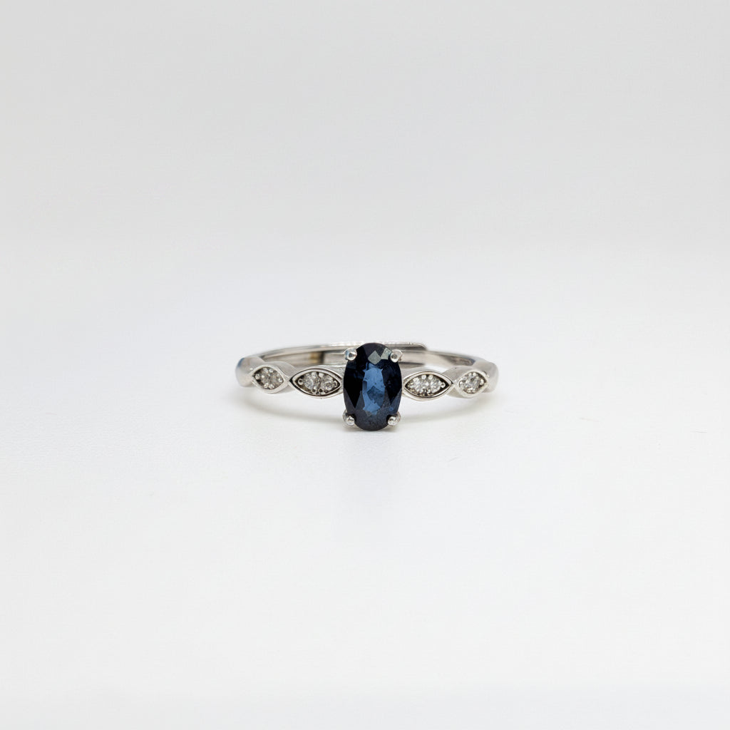 A silver ring featuring a central oval-cut dark blue gemstone and small marquise-cut side stones, displayed on a clean white surface with a soft-focus blue flower in the background.