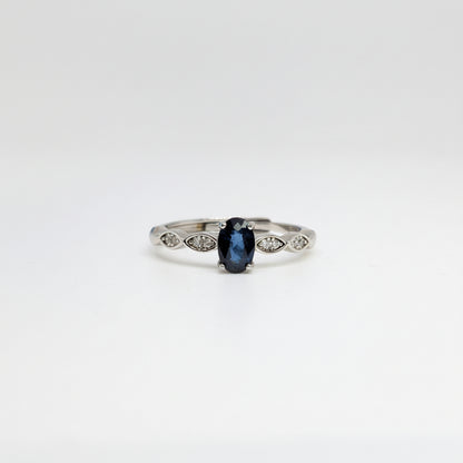 A silver ring featuring a central oval-cut dark blue gemstone and small marquise-cut side stones, displayed on a clean white surface with a soft-focus blue flower in the background.