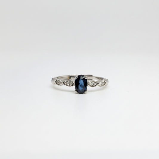 A silver ring featuring a central oval-cut dark blue gemstone and small marquise-cut side stones, displayed on a clean white surface with a soft-focus blue flower in the background.