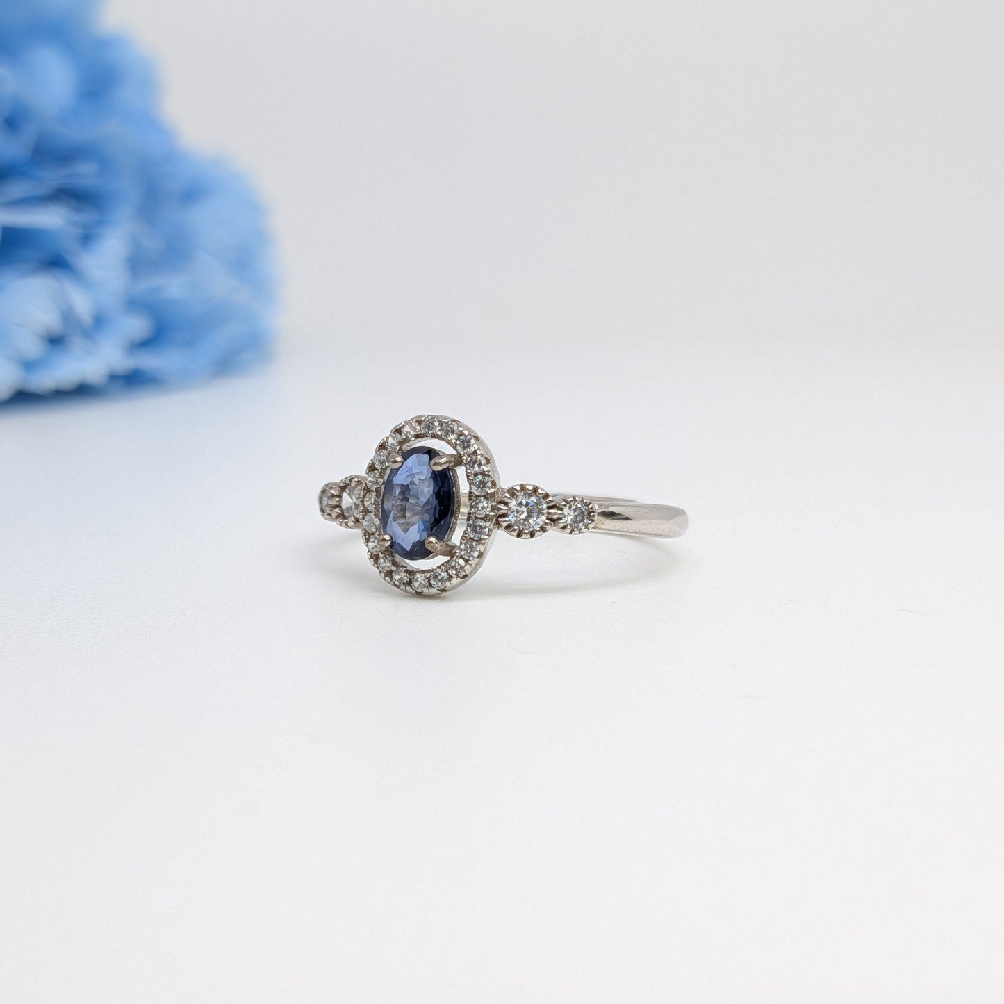 A silver-toned halo ring featuring an oval blue sapphire center stone and small surrounding clear stones, displayed on a clean white surface with a soft-focus blue flower in the background.