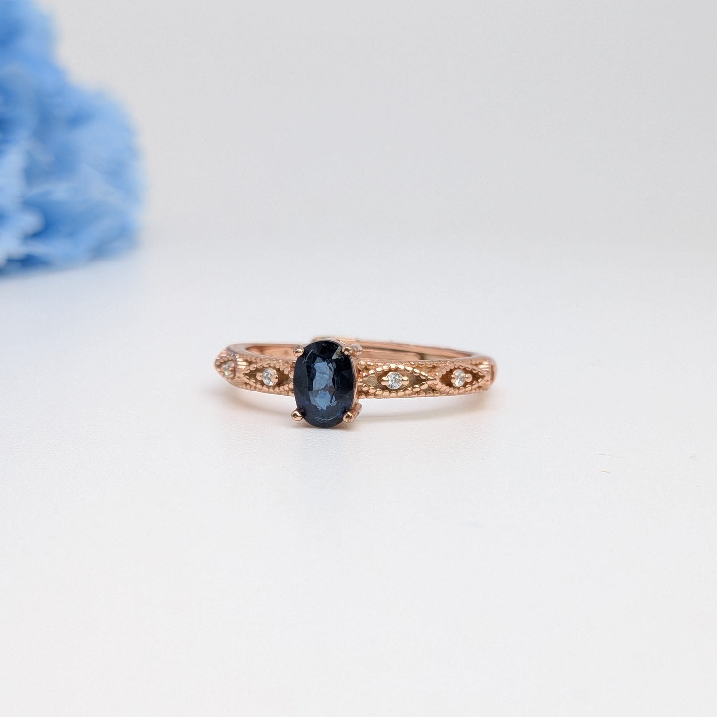 A rose gold ring with an oval dark blue gemstone and delicate filigree detailing on the band, photographed on a white background with a soft-focus blue flower in the upper left corner.