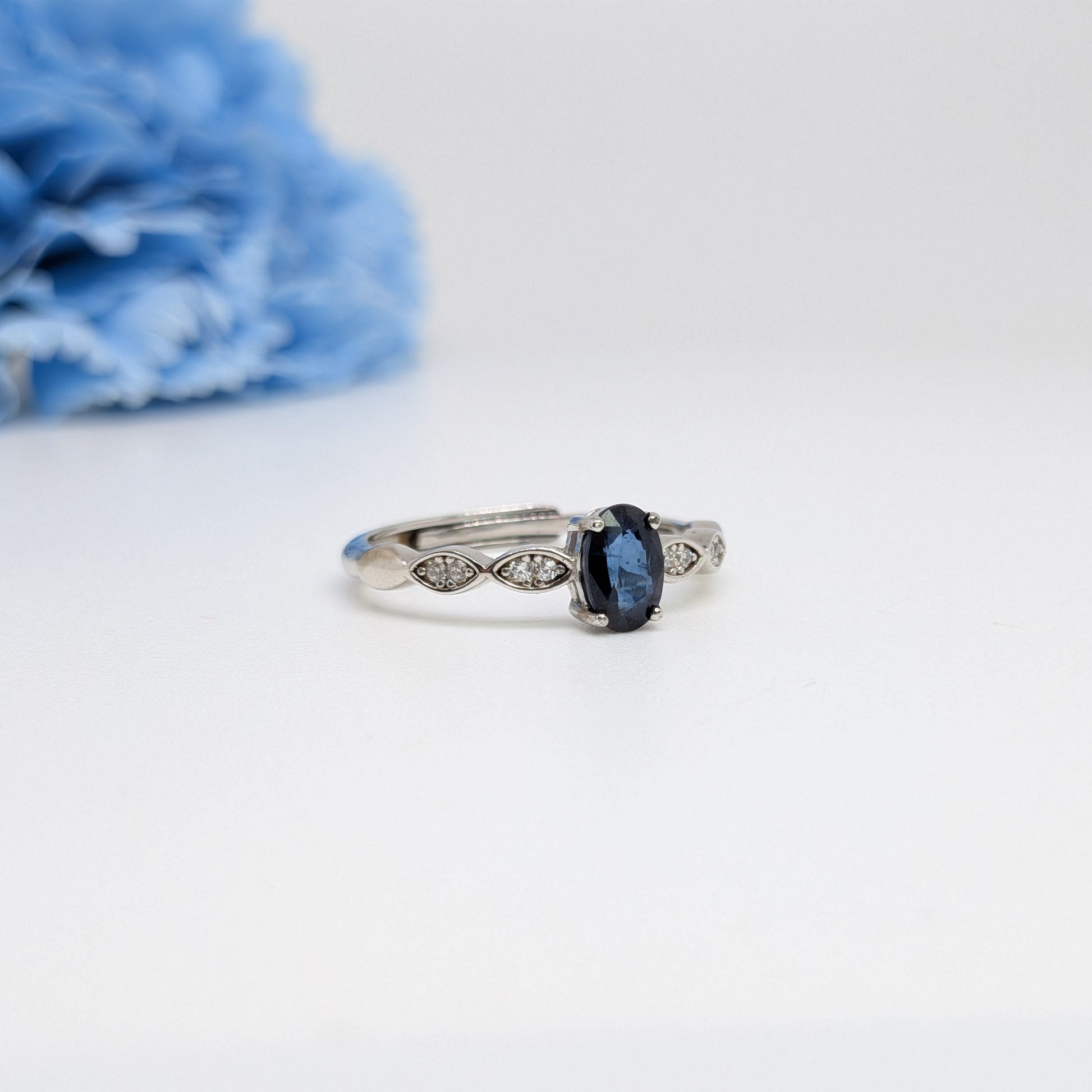 A silver ring with an oval-cut dark blue sapphire center stone and small diamond accents on the band, photographed on a white surface with a soft-focus blue flower in the background.