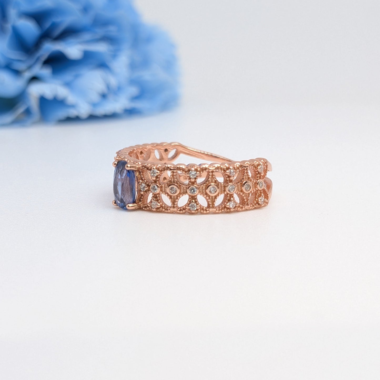 A rose gold filigree ring with a blue rectangular gemstone, displayed on a clean white surface with a blurred blue flower in the background.